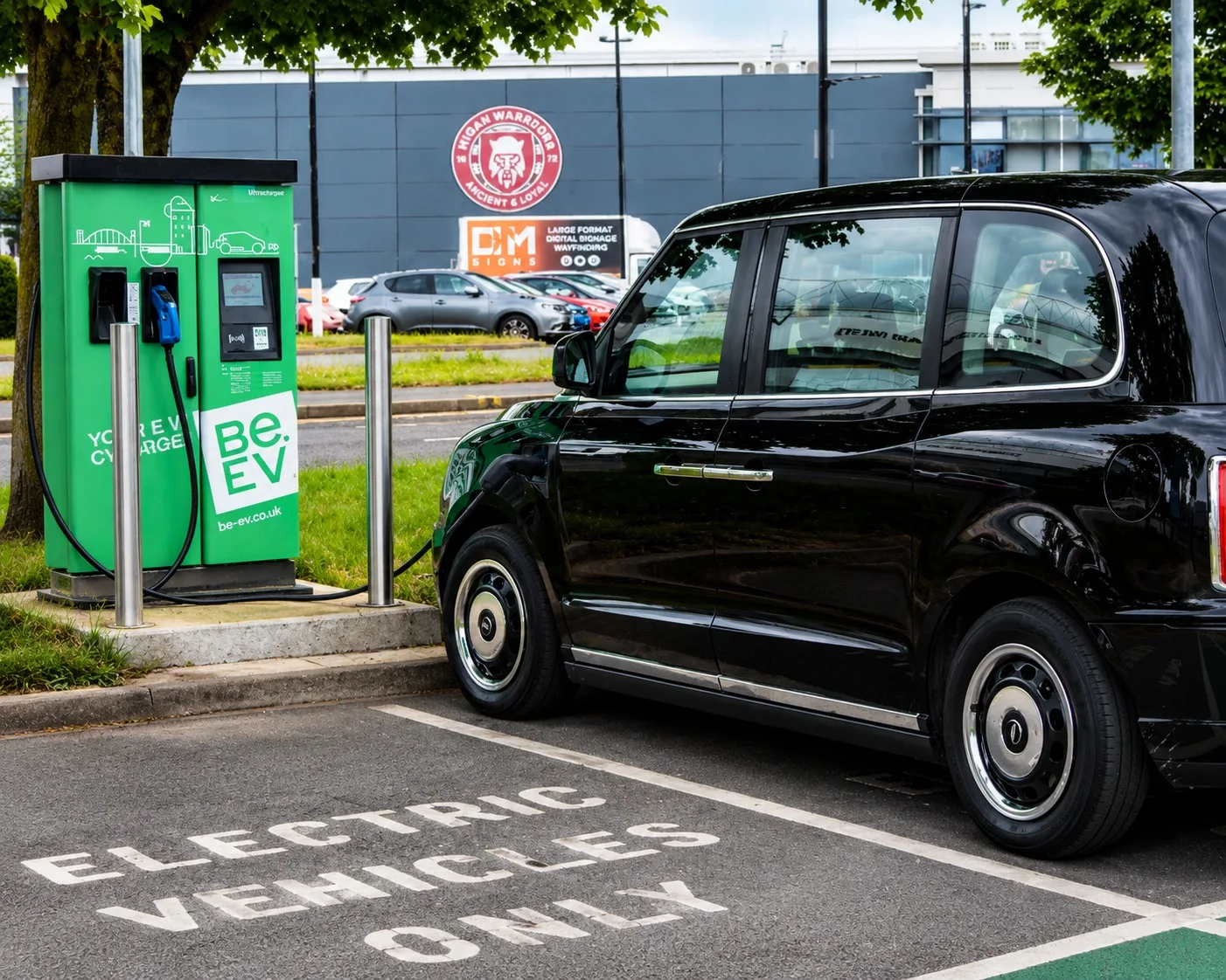 Black electric hackney carriage on charge from a green rapid charging unit, in a car park bay marked Electric Vehicles Only