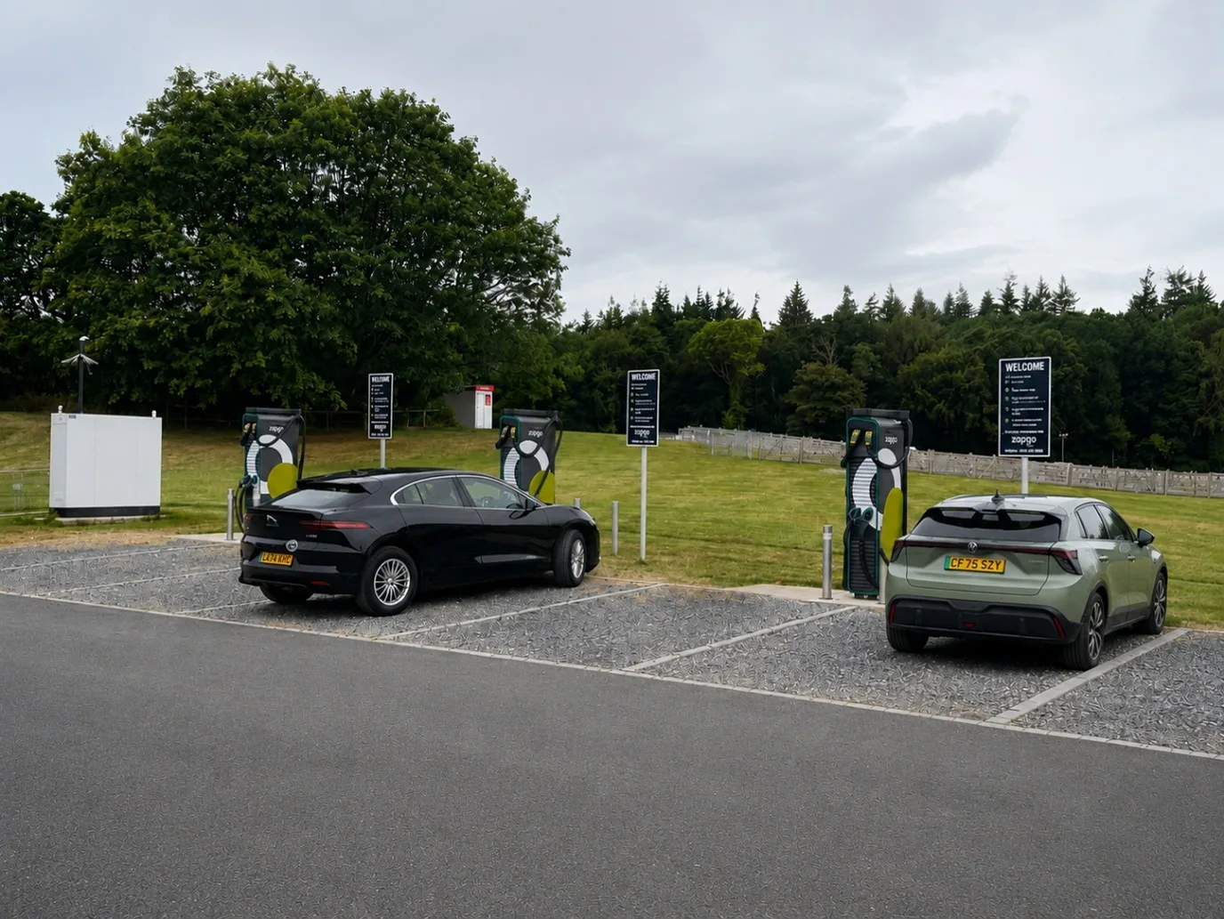 Three ultra-rapid charging units on grass-reinforced bays at the edge of a racecourse field, with two cars on charge and a ground-mounted electrical enclosure alongside