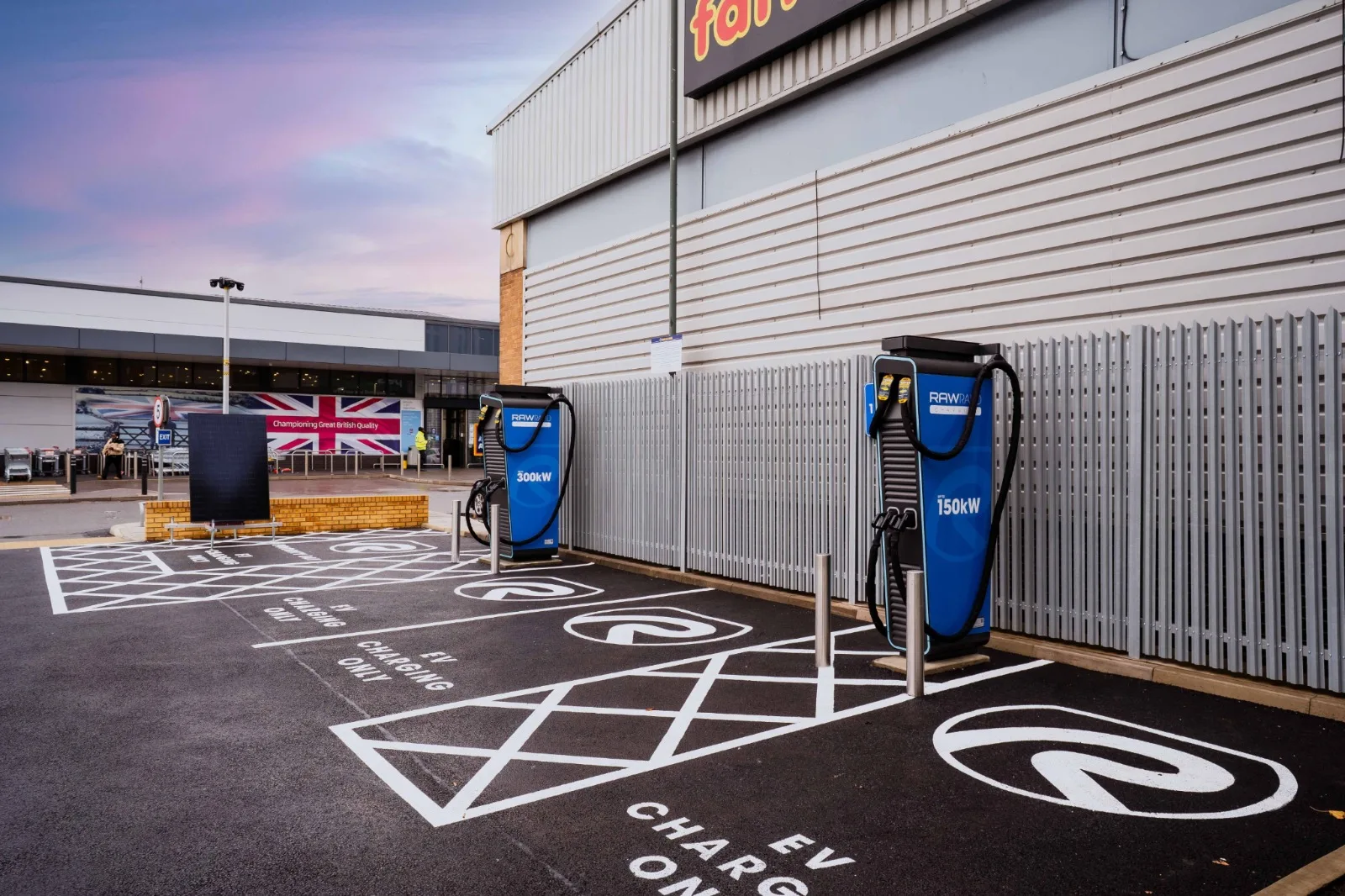 Two blue rapid charging units standing against a palisade fence at a retail park, serving freshly marked EV charging only bays outside a large store